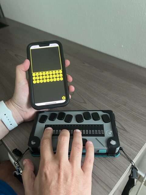 La foto presenta un iPhone con el app BrailleBuzz y una línea Braille.