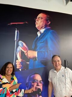 Foto de Manolo y Miltia posando junto al cartel en el concierto de Gilberto Santa Rosa en el lobby del Coliseo de Puerto Rico