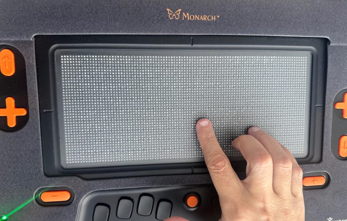 La foto muestra el dedo índice de la mano de Manolo leyendo Braille en la tableta Monarch de la APH.