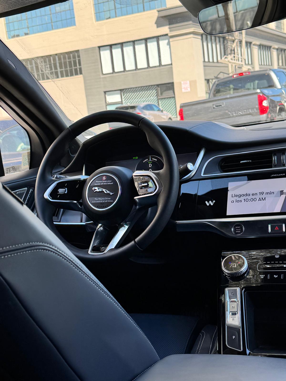 Foto que presenta los controles de la parte frontal del vehículo Waymo, incluyendo el guía sin ningún conductor. La foto es tomada dentro del vehículo desde el asiento de atrás.